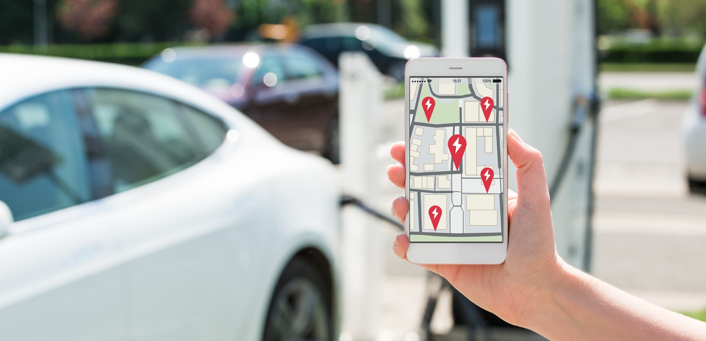 Un teléfono móvil muestra un mapa con puntos de recarga para vehículos eléctricos junto a un coche conectado a un cargador