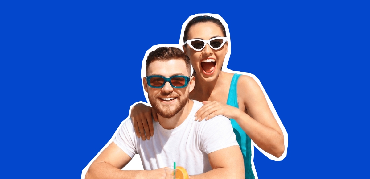 A smiling couple wearing sunglasses pose against a blue background, conveying a cheerful, relaxed and summery mood.